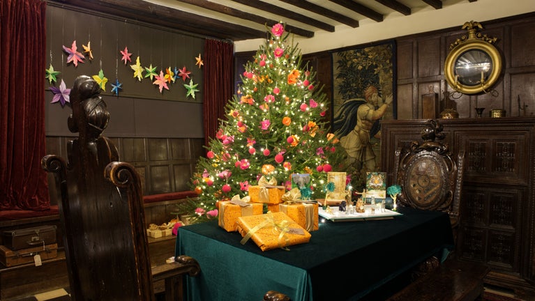 Historic room decorated for Christmas with a glowing tree, colourful ornaments, and wrapped gifts on a green-covered table.